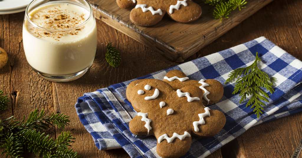 Galletas de jengibre con glaseado blanco y un vaso de ponche de huevo sobre una mesa rústica con decoraciones navideñas.